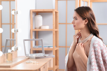 Young woman trying on elegant pearl necklace at dressing table indoors - obrazy, fototapety, plakaty