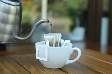Pouring hot water into cup with drip coffee bag from kettle on wooden table, closeup