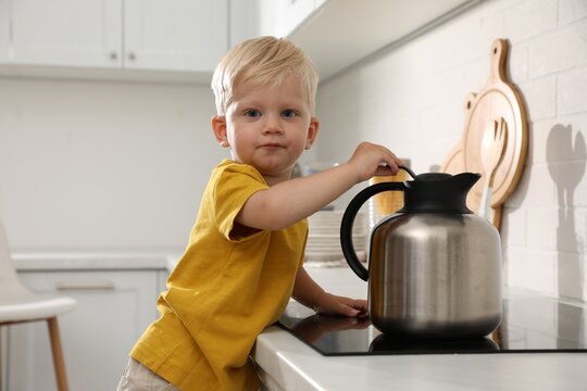 Curious little boy playing with kettle on electric stove in kitchen