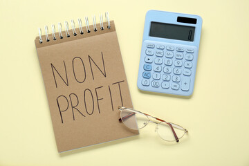 Notebook with phrase Non Profit, glasses and calculator on beige background, flat lay