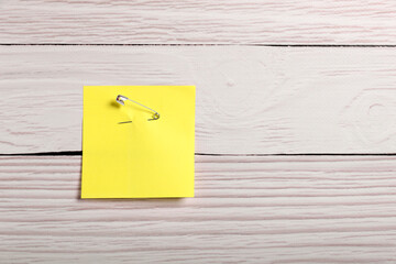 Yellow paper note attached with safety pin to white wooden background, top view. Space for text