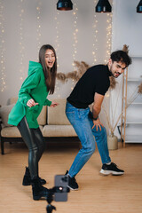 Fototapeta premium Funny man and woman recording video for social media dancing in front of camera, using cell phone on tripod, home interior. Video live streaming.