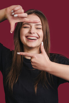 Portrait of young beautiful woman cheerfuly smiling making a camera frame with fingers isolated over cherry-colored background.