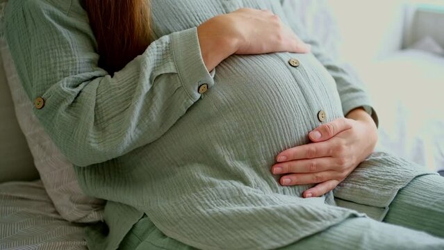 Unrecognizable brunette pregnant woman caressing,stroking touching big belly, baby bump sitting, lying on bed, close-up, cropped shot.Happy natural, calm pregnancy.Having rest, relaxing girl at home.