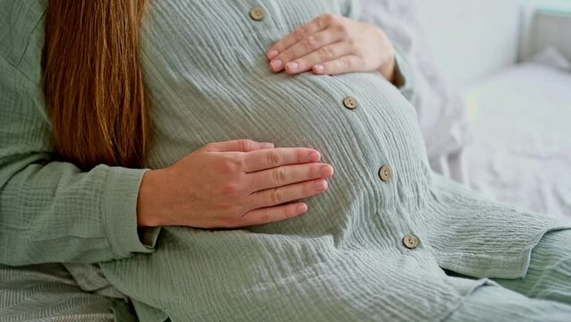 Unrecognizable brunette pregnant woman caressing,stroking big belly, baby bump sitting, lying on bed, close-up, cropped shot.Happy natural, calm pregnancy.Having rest, relaxing girl at home. 