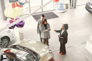 Black saleswoman standing at auto showroom with young couple