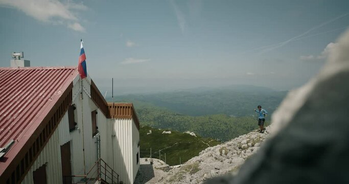 Hiker walkig from behind the rock towards the cottage on mountain Snežnik. Small fluter of the Slovenian flah on top of the roof. Green karst in background.