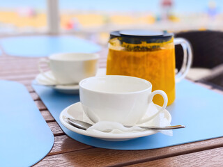 Two white cups and a transparent teapot with sea buckthorn tea on a blue background in an outdoor cafe. Beach, summer. Mobile photography. Horizontal.