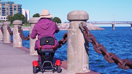 Woman with disability in the wheelchair mobility scooter enjoying park near lake water. Carefree woman with mobility issue uses electric three wheel wheelchair scooter having fun.