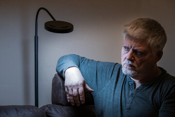 An elderly man sits lonely and depressed in a room. He looks tired. A floor lamp illuminates his face.