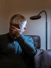 Fototapeta premium An elderly man sits lonely and sad in a room. He looks depressed. A lamp illuminates his face.