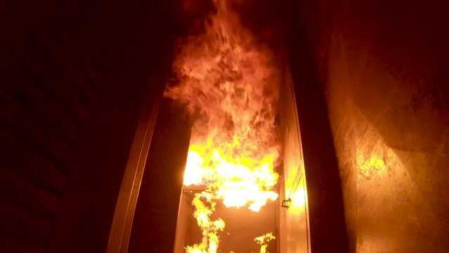 Fire ignites in burn building and flashover on ceiling sends flames rolling down hallway toward camera. Firefighter training. Tilt up.