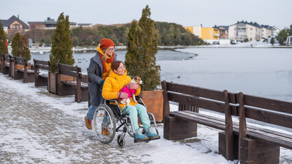 A woman in a wheelchair walks with her friend and a dog by the lake in winter.