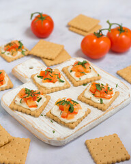 Quick cooked appetizer of cream cheese on a cracker with tomato and herbs on a wooden board and a light background, in the background are tomatoes and a cracker, in the foreground a cracker