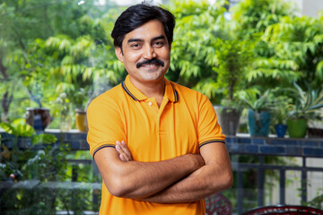 Portrait of young happy Indian mustache man wearing orange t-shirt standing cross arms with a smile on his face at home.