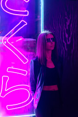 Fototapeta na wymiar Defocused beautiful young woman in cosmic glasses standing next to colorful neon light at night with selective focus. Party disco neon nightclub vibes