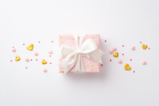 St Valentine's Day concept. Top view photo of big pastel pink present box with silk ribbon bow golden hearts and sprinkles on isolated white background