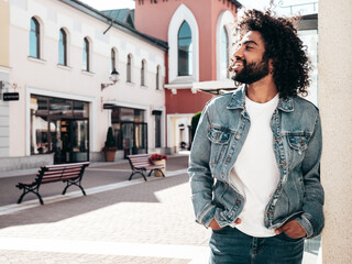 Handsome smiling hipster  model. Sexy unshaven Arabian man dressed in summer jeans jacket clothes. Fashion male with long curly hairstyle posing in street at sunset. Cheerful and happy