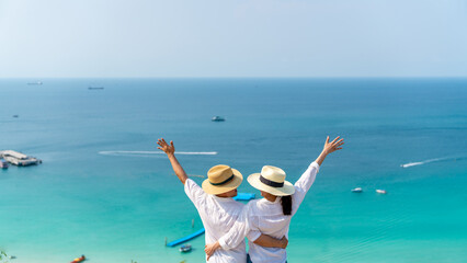 Happy Asian family couple enjoy outdoor lifestyle travel tropical island on summer holiday vacation. Husband and wife holding and raise hands up at mountain peak and looking to blue ocean in sunny day