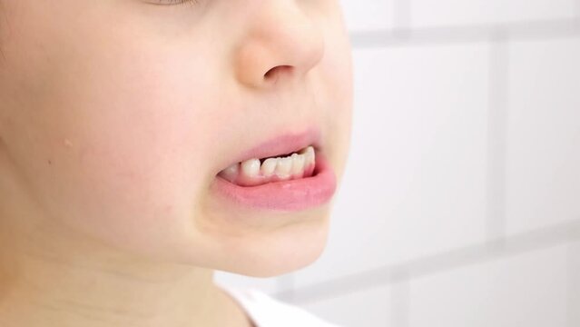 The child moves a loose milk tooth and presses on it with a finger. Open mouth of a caucasian 6 year old kid in a white t-shirt on a bathroom background. Lower cutter. Healthcare. Part of the body.