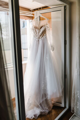 Fashionable beautiful lace wedding dress hanging on hanger in hotel room. morning preparation wedding concept. vintage wedding gown.