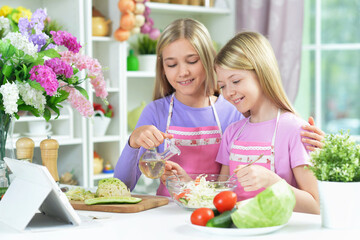 Two girls in pink aprons preparing salad on kitchen with tablet