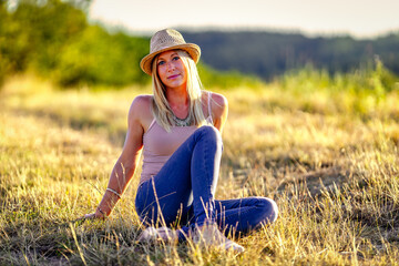 Woman with straw hat blond with long hair sits in a meadow at sunset..