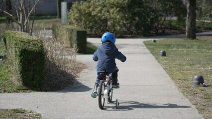 Back of child riding bicycle outdoors wearing helmet. One little boy rides bike outside at park pathway. Kid cyclist