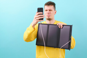 A man holds a foldable portable solar panel and suspiciously looks at the phone on blue background. Equipment. Communication. Connect. Electronics. Engineering. Photovoltaic. Generation