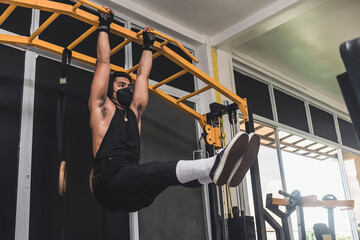 Fototapeta na wymiar An asian man wearing a face mask doing hanging leg raises at the workout station at the gym. Training abs and core strength.