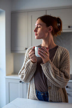 Young Woman Relaxing At Home In Kitchen Holding Hot Drink With Eyes Closed