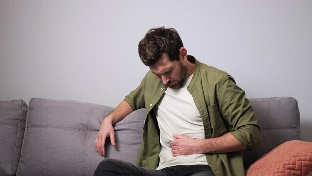 Terrible pain in the stomach. A young man in casual clothes suffering from abdominal pain, holding his stomach, sits on the sofa at home. Man suddenly feeling strong stomach ache, gastritis problem.