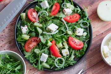 Fresh spring salad with arugula, feta cheese, red onion and tomatoes in a black bowl. Recipe ingredients on chalkboard background.