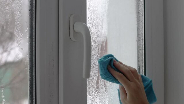 A womans hand wipes the condensate from the window glass with a towel.