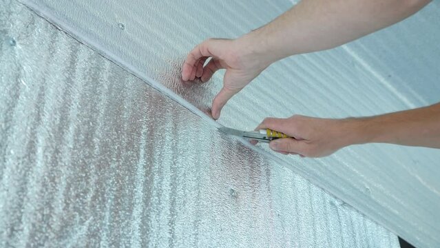 Construction roll heat-insulating material. A man cuts the vapor barrier insulation on the wall with a construction knife. Covered with a thin layer of aluminum.