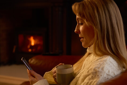 Woman At Home In Lounge With Cosy Fire Using Mobile Phone