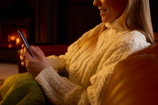 Close Up Of Woman At Home In Lounge With Cosy Fire Using Mobile Phone
