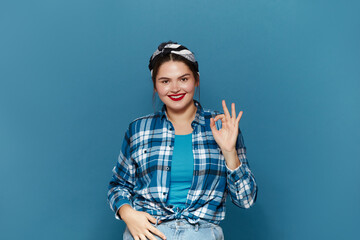Smiling Woman Ok Gesture Isolated. Portrait of Satisfied Plus Size Girl Standing, Looking at Camera and Showing Ok Sign Gesture. Indoor Studio Shot Isolated on Blue Background  - obrazy, fototapety, plakaty