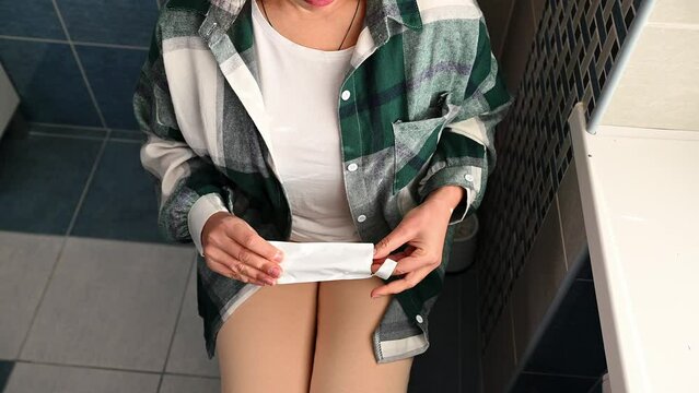 Top view of a probably pregnant woman, sitting on toilet bowl in the home bathroom, unpacking and removing a pregnancy test from package, anxiously holding it in her hands, before checking pregnancy