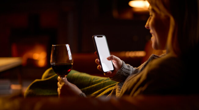 Woman At Home Lying On Sofa In Lounge With Cosy Fire With Blank Screen Mobile Phone Holding Glass Of Wine