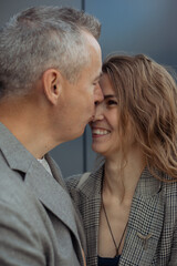 Vertical romantic couple of mature gray haired man kissing to nose fair haired laughing woman against gray wall. Family