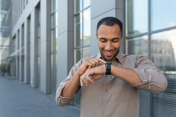 Latin American businessman in shirt cheerful talking with friends using smartwatch, man outside office building using gadget for artificial intelligence commands.