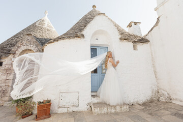 Side view of young woman bride with long flying bridal veil, standing near blue door of old white...