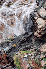 Alpine flowing waterfall washes various color karst overlays of granite rocks, scarce multi-colored rock vegetation nestled in open areas of wet rock, vertical shot, Aosta valley, Italy