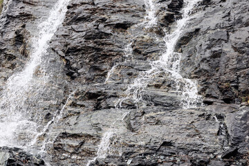 Сlose-up monochrome shot of flowing and foaming waters of small alpine cascade over gray karst layers of granite, Aosta Valley, Italy