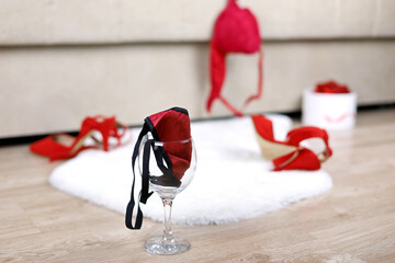 Wine glass with female thong panties, removed red bra and shoes on a floor near bed. Sex concept,...