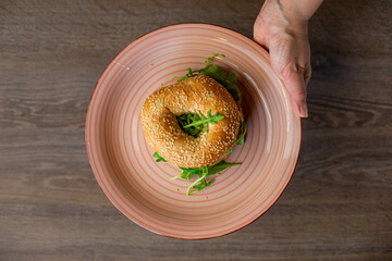 Cropped photo of woman holding plate with fresh appetizing burger with hole in bun, arugula on wooden table. Cooking.