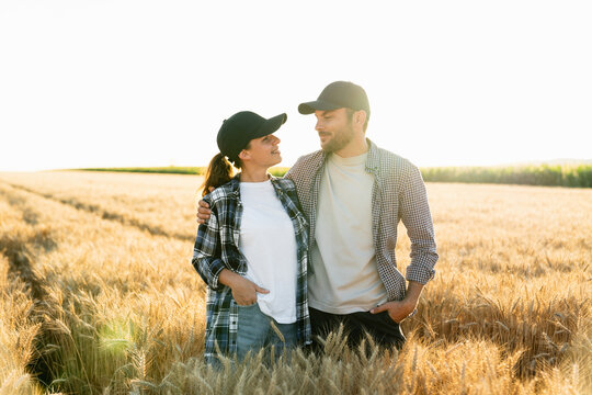 A couple of farmers in plaid shirts and caps stand embracing on agricultural field of wheat at sunset