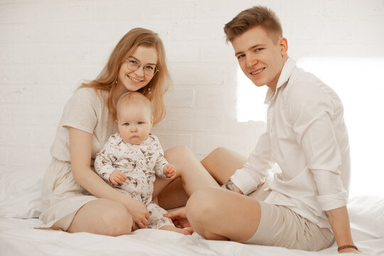 Portrait of happy family of three on white background. Mother, father and infant baby sit on bed and hug. Photo of young couple and child in white family look. Parental affection, childhood, love