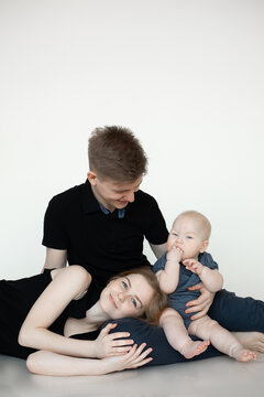 Young family from three in black looks on white background. Dad sit on floor and hold infant baby licking hands, joyful mom lie on leg of husband. Family photo, parenting, firstborn, love, feelings